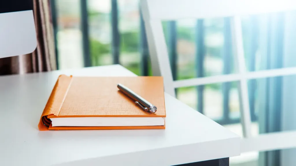 Un cuaderno de papel kraft cerrado junto a un bolígrafo sobre un fondo blanco y minimalista