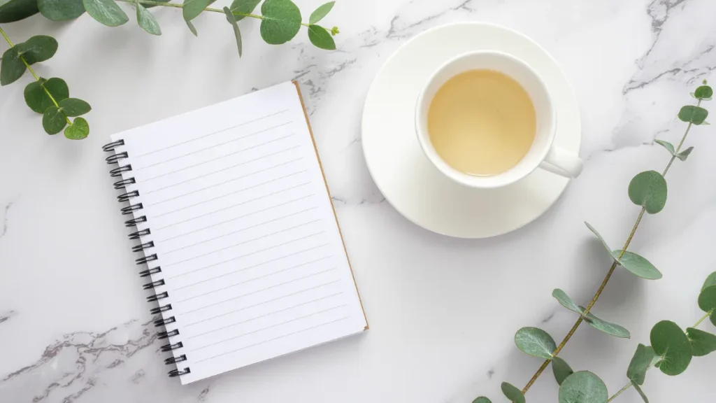 Escritorio minimalista con un cuaderno en blanco, una taza de té y hojas verdes, representando el crecimiento personal y bienestar de Mejor que Ayer.