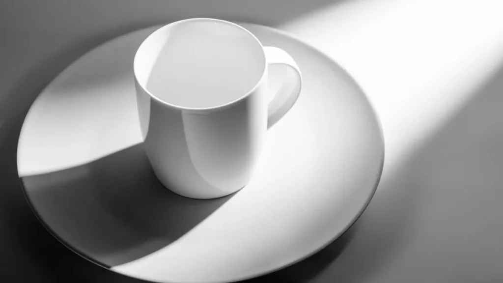 Una taza de cerámica blanca vacía sobre una superficie gris con sombras marcadas de luz natural